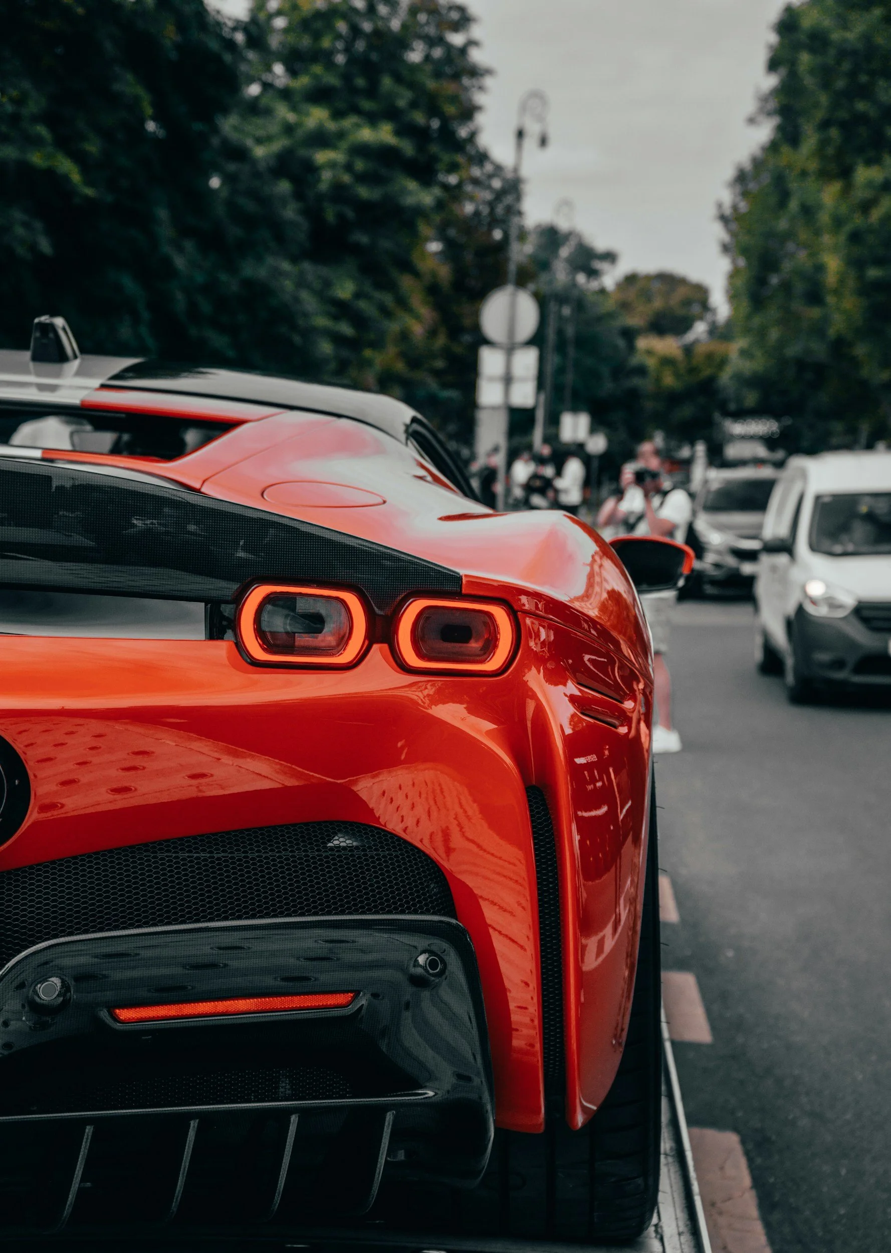 Close-up of a red sports car with modern taillights and a rear diffuser, parked on a street with trees and people in the background.