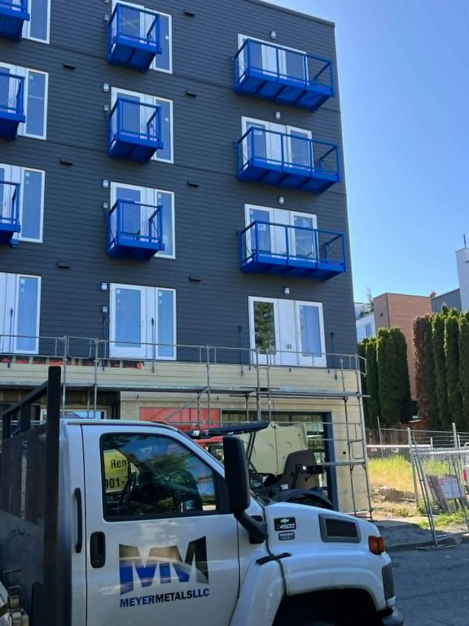 A multi-story residential building with blue balconies and large windows. Construction work is ongoing at the building's lower level, with scaffolding and a white utility truck labeled 'MEYERMETALSLLC' parked in front.