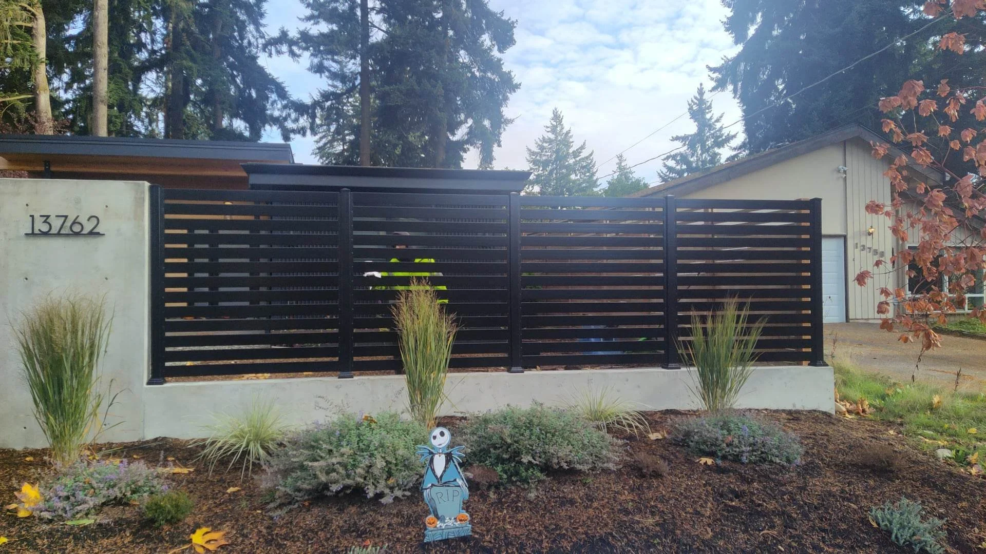 A modern house with a tall black metal horizontal slat fence, numbered 13762, surrounded by a landscaped garden with various plants, including tall grasses and flowering ground cover. The house has beige siding, a white garage door, and is set agains