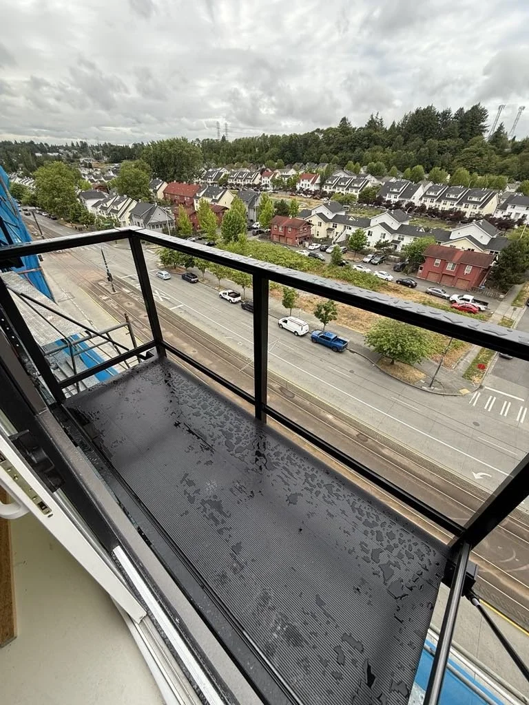 View from a high-rise apartment balcony showing city streets, parked cars, a row of houses, and trees under a cloudy sky.