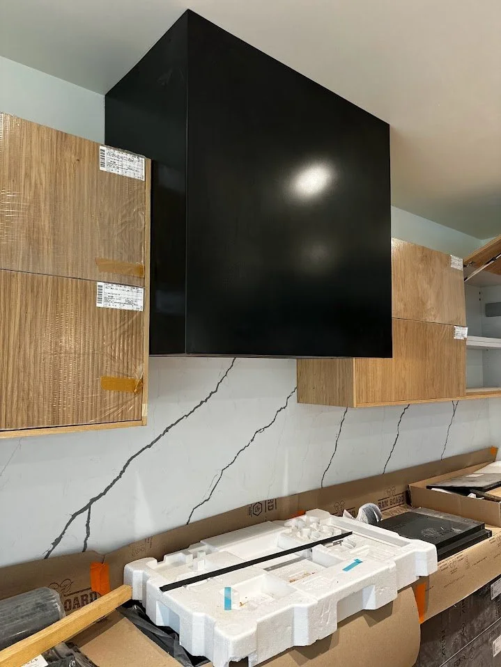 Black kitchen range hood above the kitchen countertop, with wooden cabinets on either side, and a white marble backsplash with gray veining.