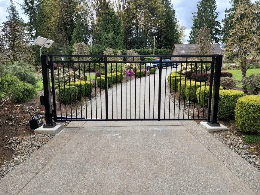 A black metal driveway gate in a residential yard, with a driveway and landscaped garden with bushes and trees behind it.