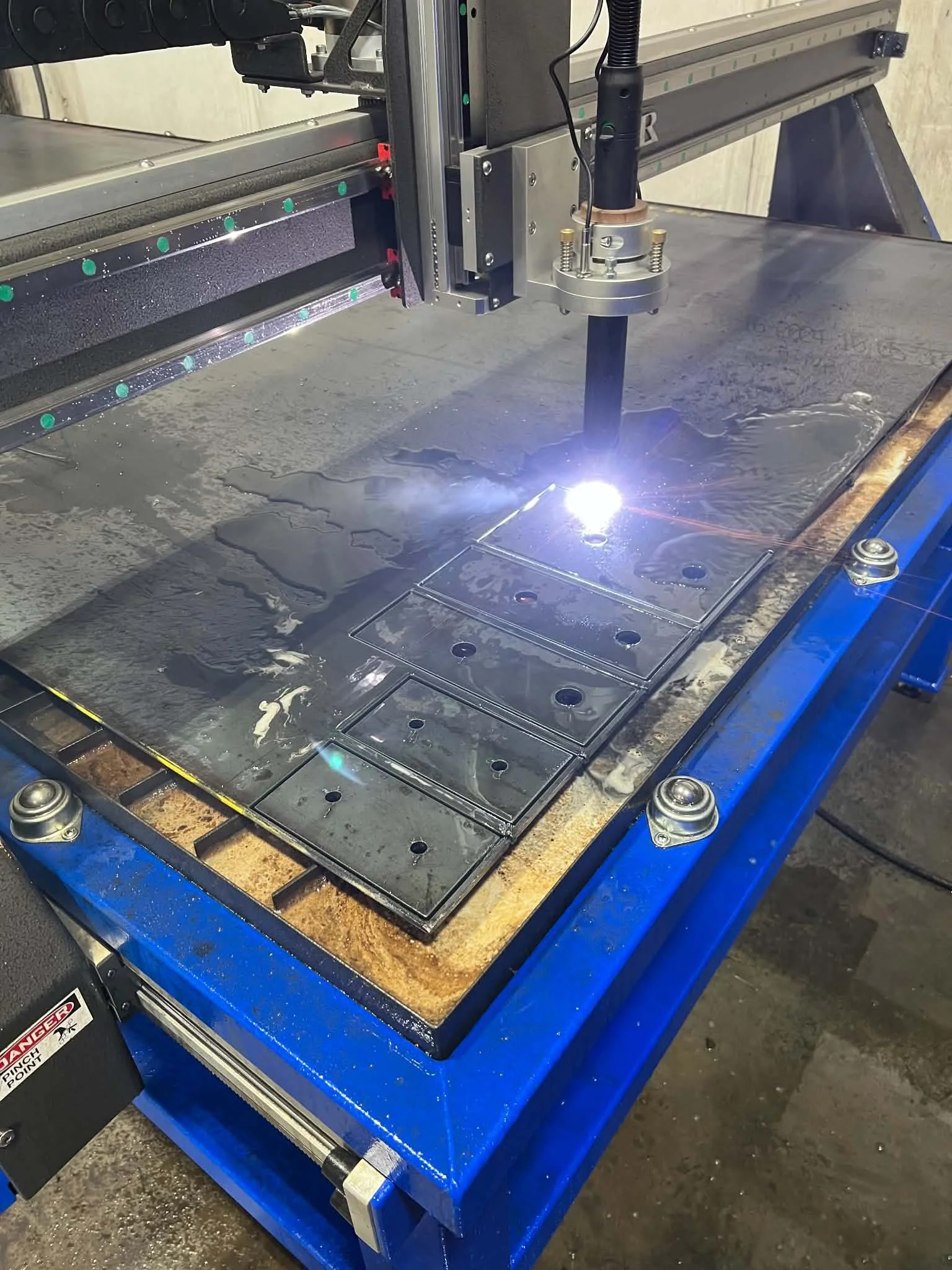 A CNC plasma cutting machine in operation, cutting through a metal sheet with bright sparks and sparks flying from the cutting torch, placed on a worktable in an industrial workshop.