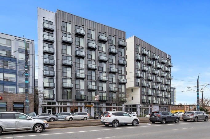 Modern multi-story apartment building with numerous small balconies, situated along a busy street with cars parked and driving by under a clear blue sky.