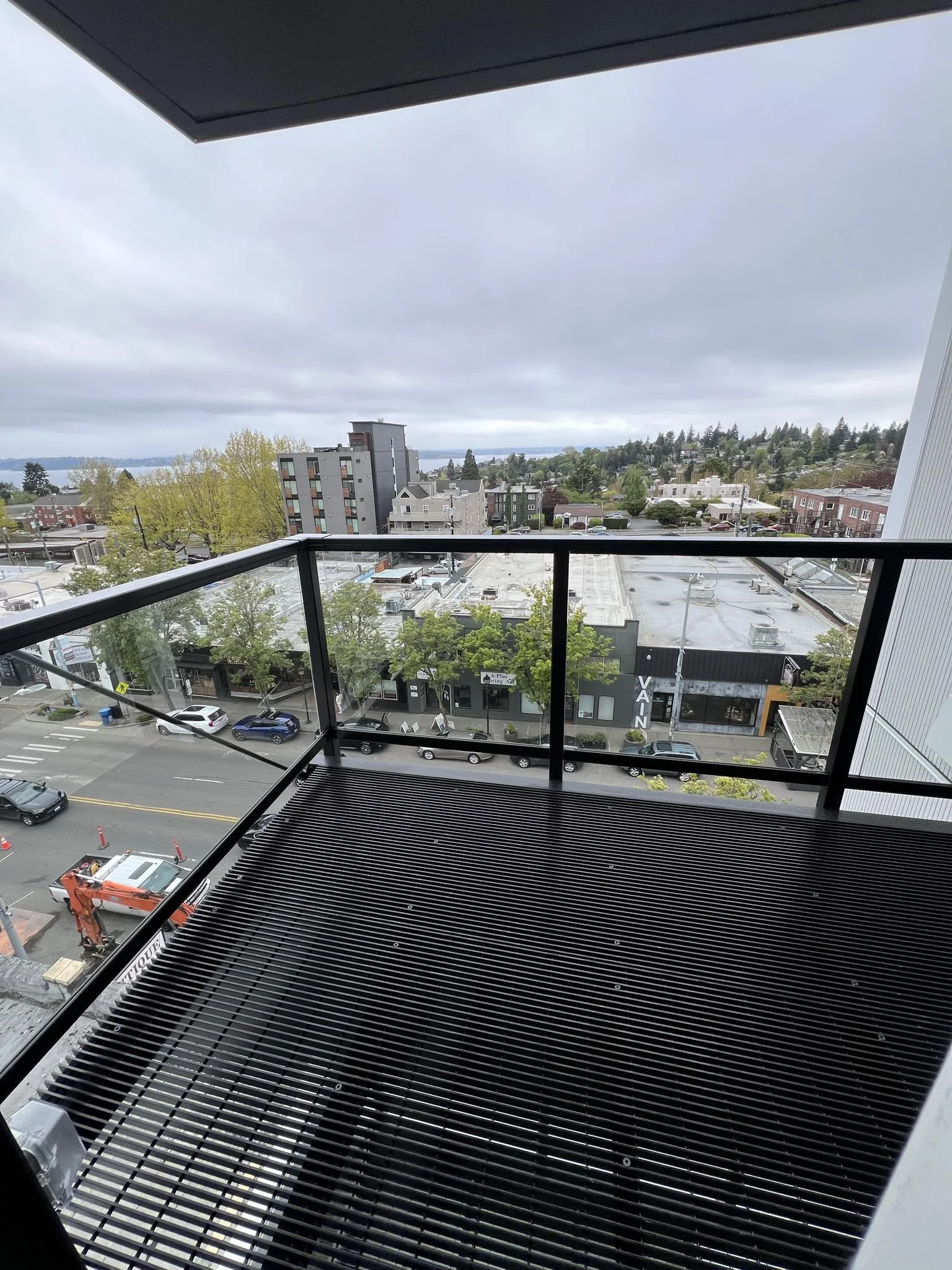 View from a balcony overlooking a city with buildings, trees, parking lots, and a body of water in the distance under an overcast sky.