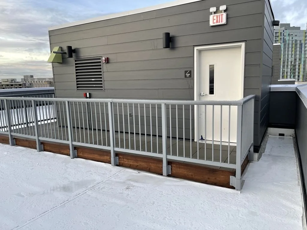 Rooftop terrace with white safety railing, gray building wall, door, exit sign, and city skyline in the background.