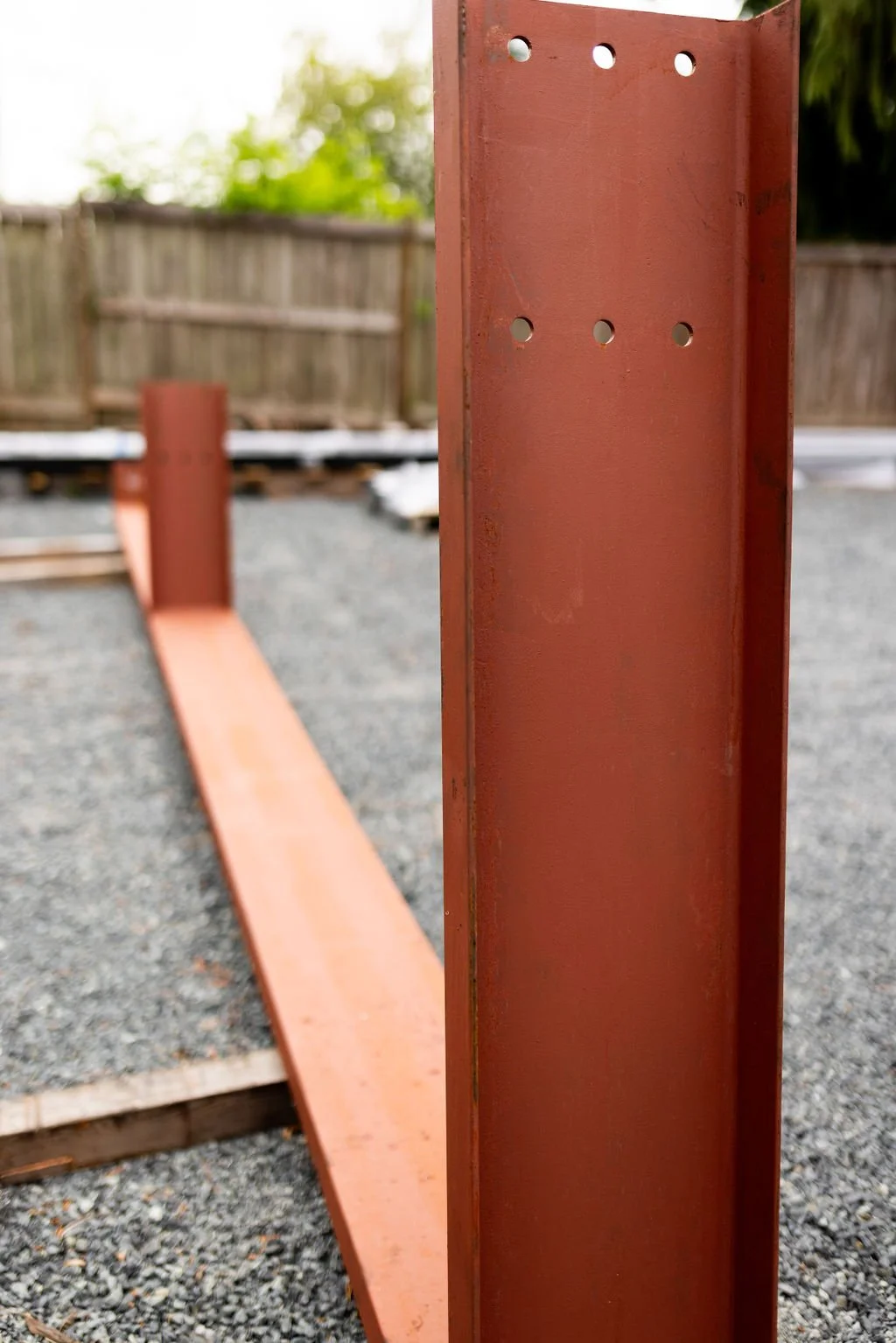 Close-up of a vertical red metal post with holes, part of a structure under construction, with a gravel ground and wooden fence in the background.