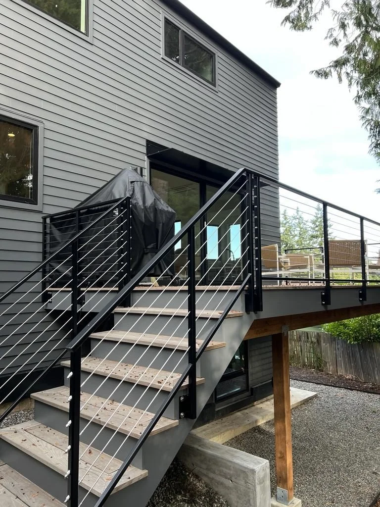 Exterior view of a modern house with gray siding, wooden stairs leading to a deck, and black metal railing.