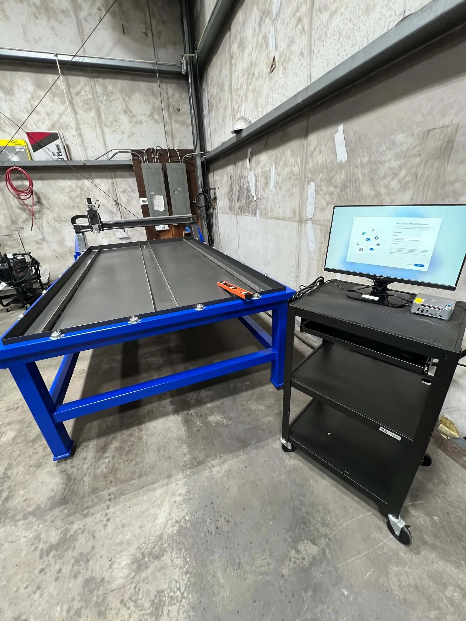 A blue metal frame table in a warehouse or industrial space with a computer monitor on a black rolling cart nearby and equipment on the wall behind.