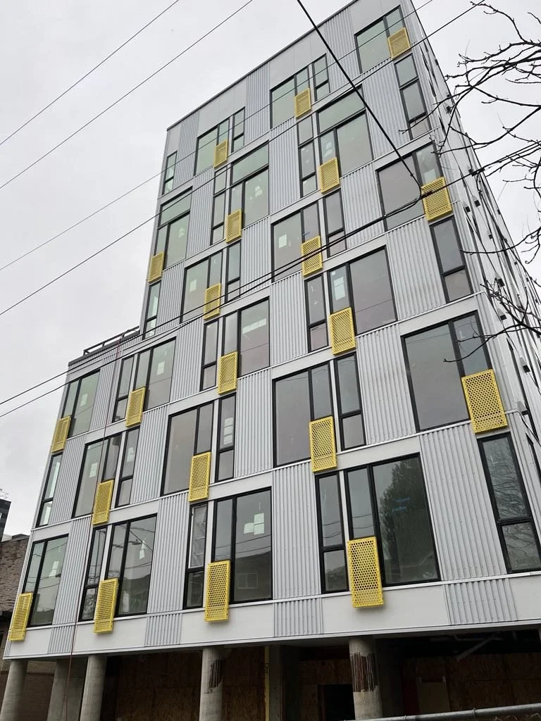 Modern multi-story building with large windows and yellow ventilation panels, with power lines overhead and a tree branch to the right.