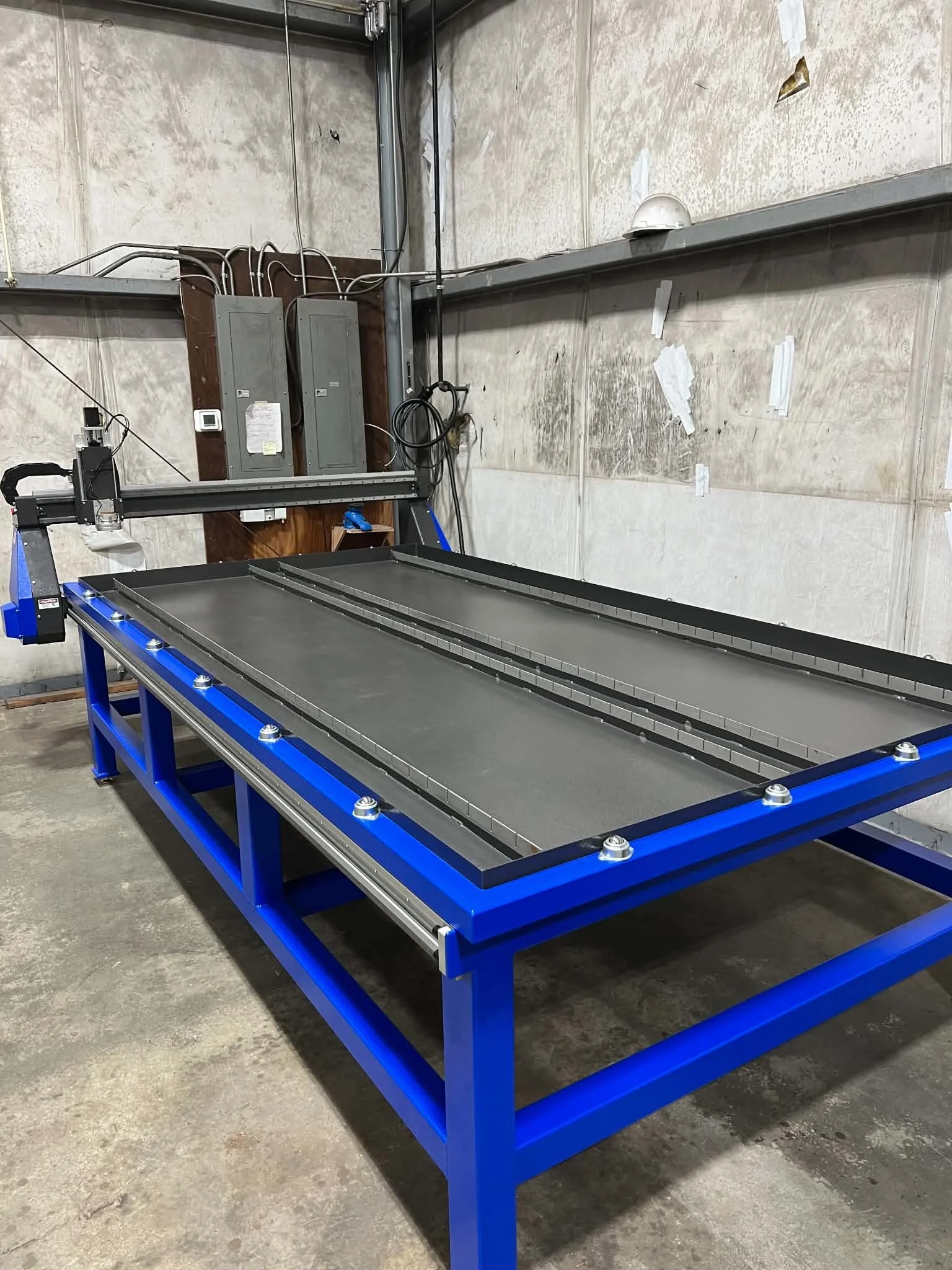 An industrial workspace with a large blue metal frame table in the foreground with a flat black surface. In the background, there are electrical components and wiring mounted on a wood panel attached to a concrete wall.