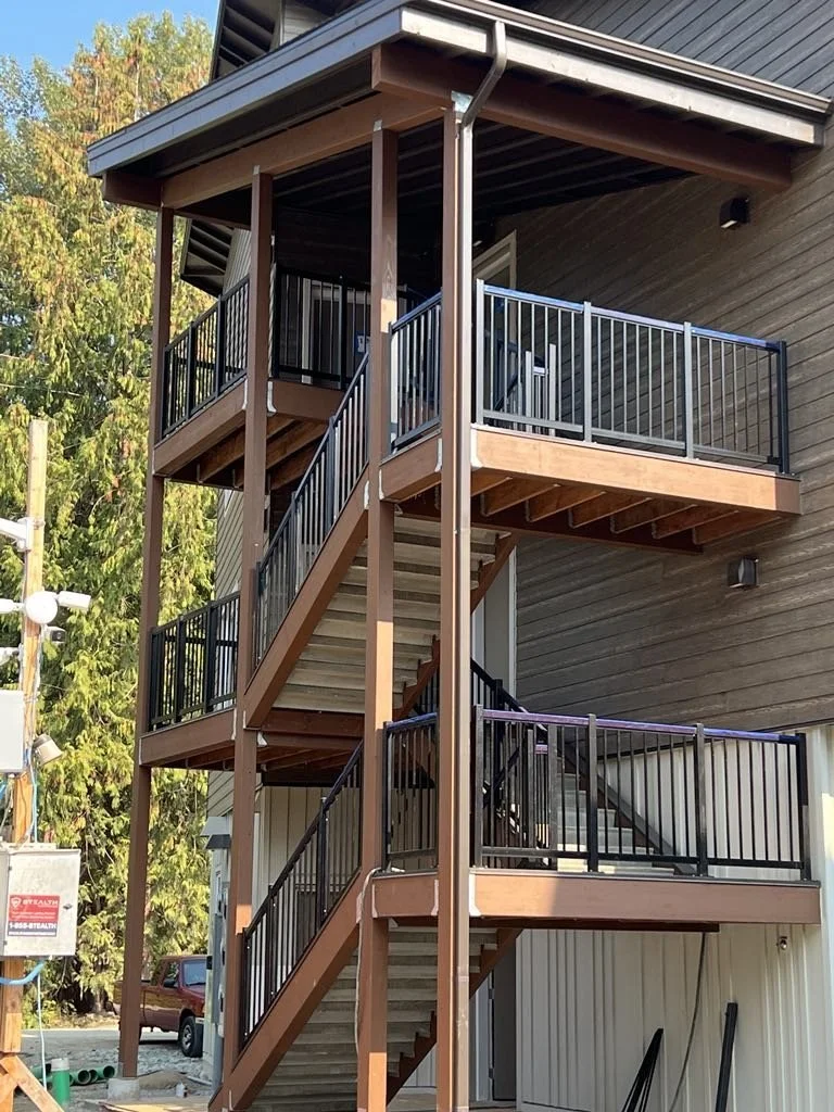 Exterior view of a three-story wooden staircase with metal railings, leading to multiple balconies on a residential building, with trees and a utility pole in the background.