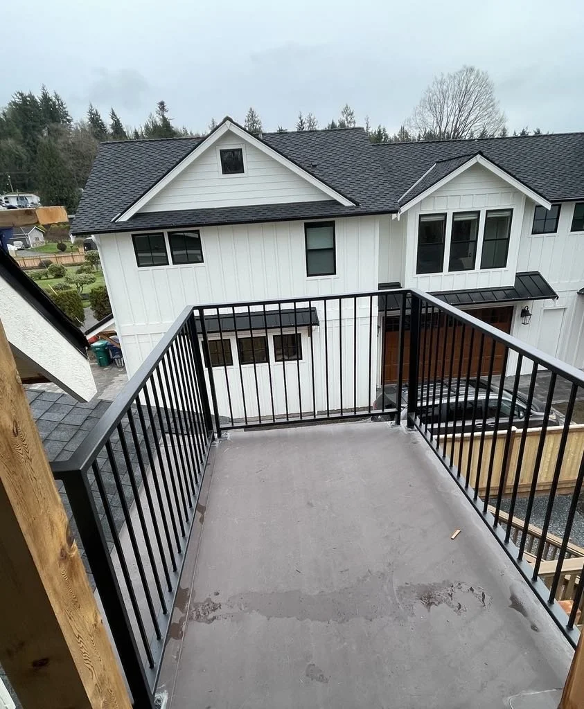 View of a new balcony with black metal railings on the second floor of a modern white house, overlooking neighboring houses and a driveway.