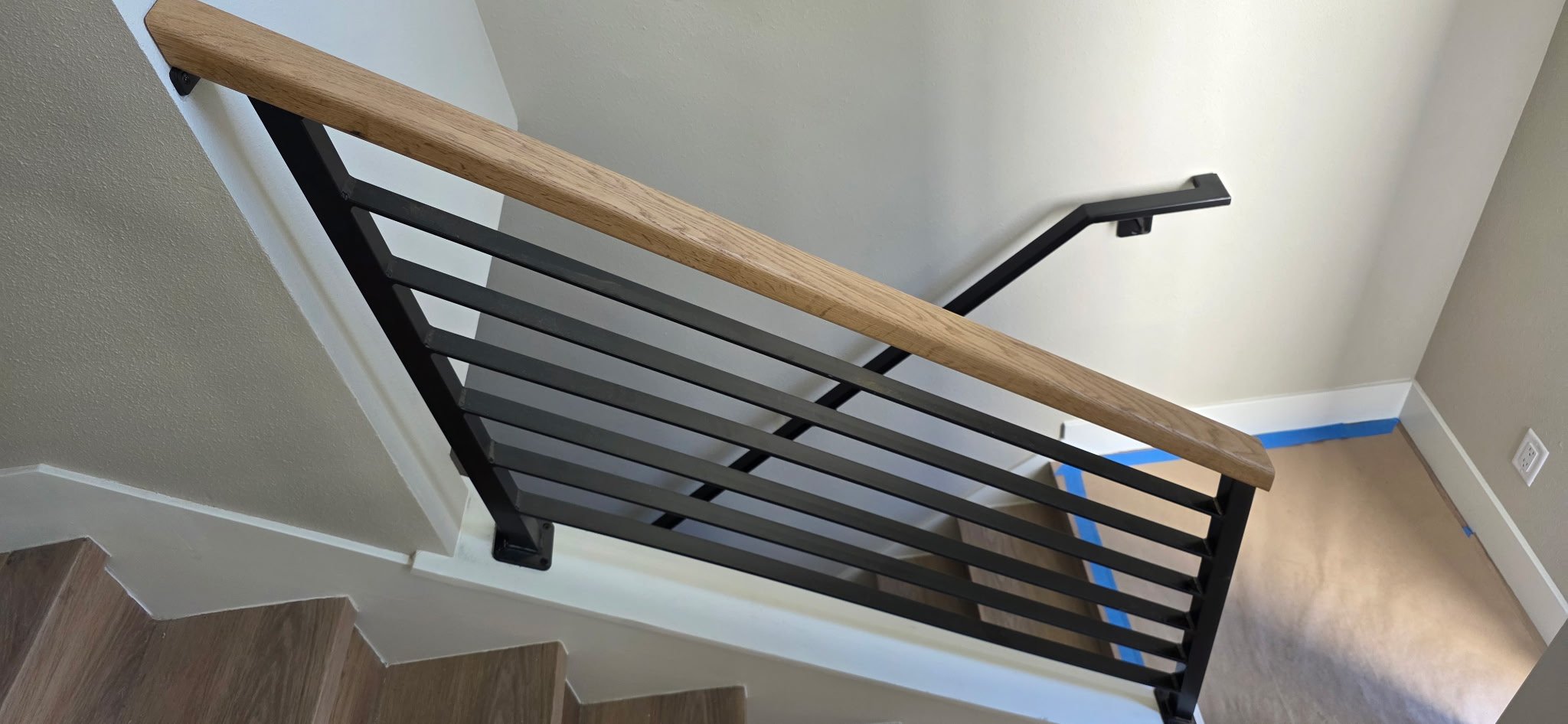 Interior view of a staircase with wooden steps, a black metal railing, and a wooden handrail. The wall adjacent to the staircase is painted light beige, and the floor is light wood. There is a blue painter's tape along the edge of the wall near the c