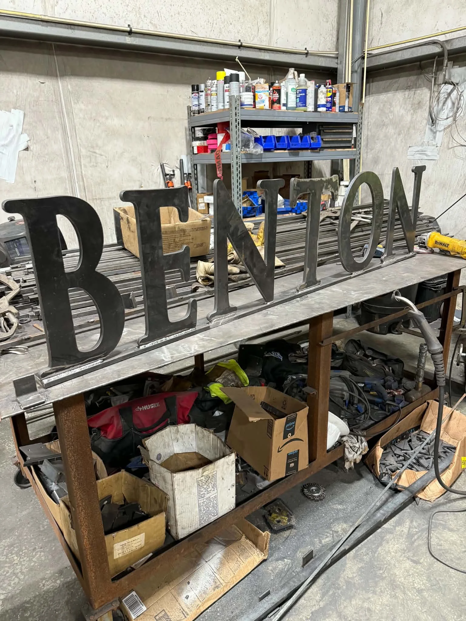A cluttered workbench in a garage or workshop with metal letters spelling 'BEYOND' on top, surrounded by various tools, boxes, and supplies, with shelves full of cans, bottles, and organizational bins in the background.