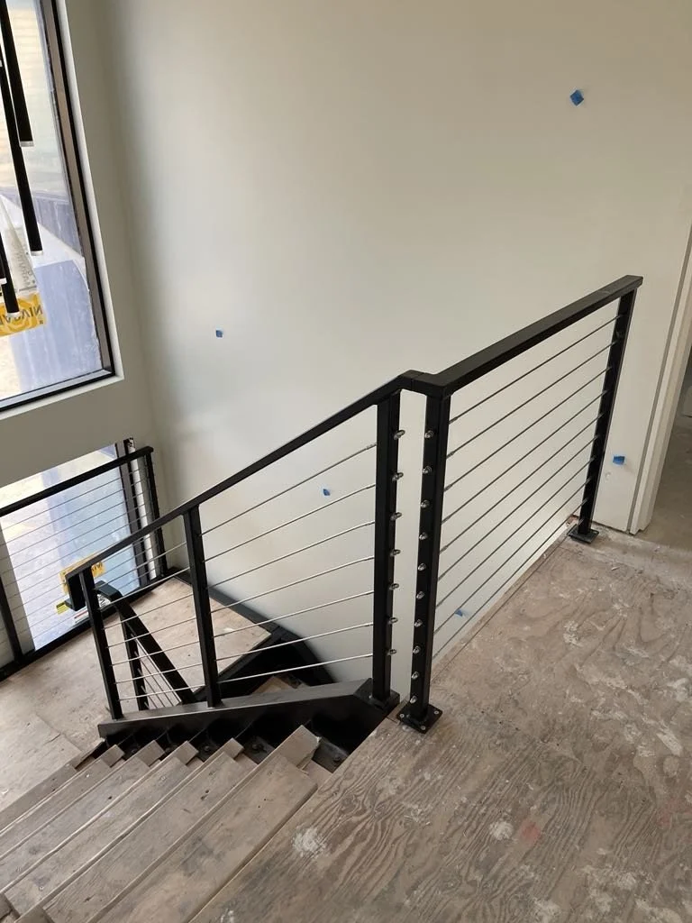 A staircase with wooden steps and a black metal handrail with cable railing, descending to a lower level beside a window in a partly finished building.