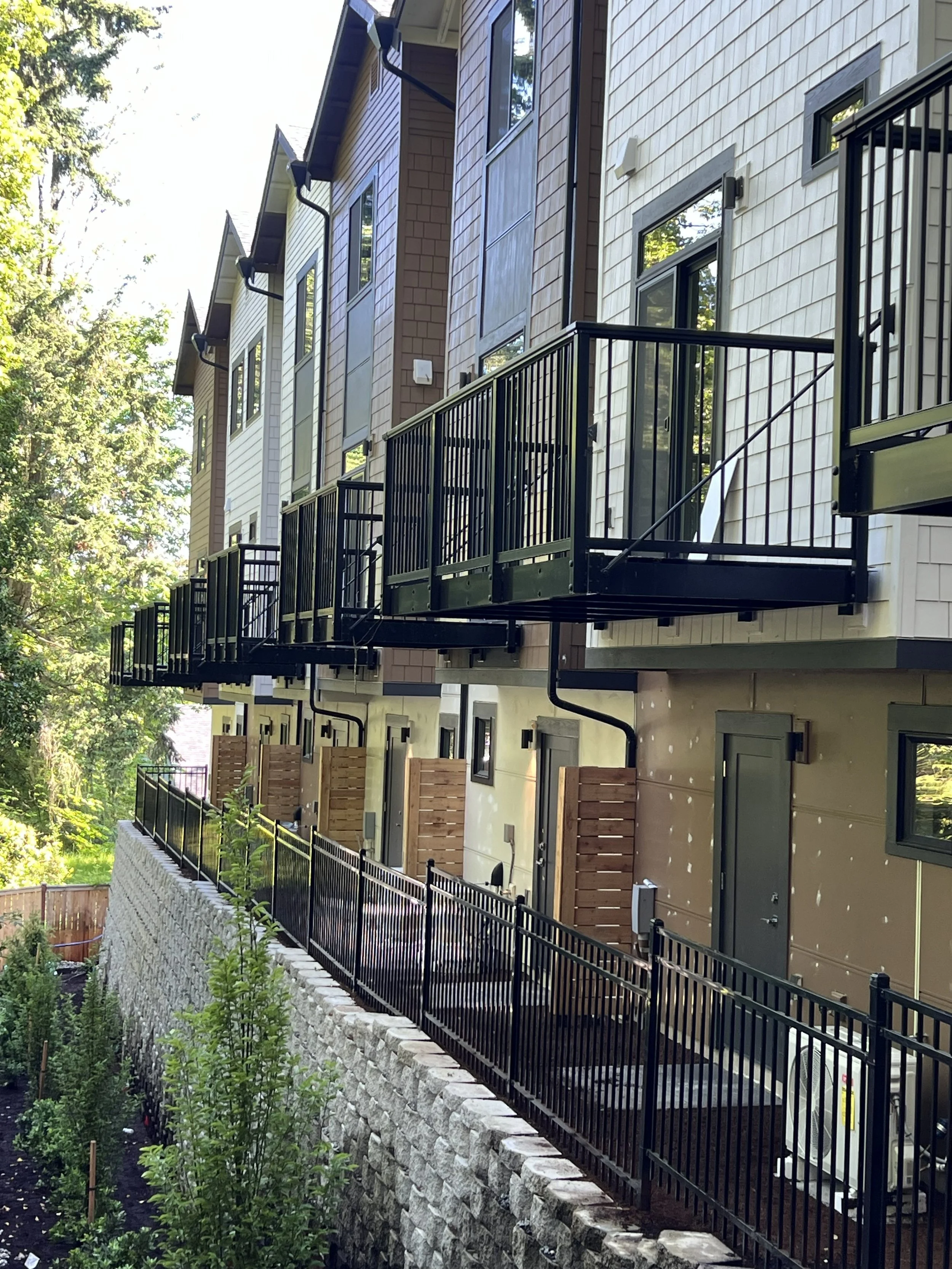 Multiple new residential buildings with private balconies, exterior walls with beige and brown siding, black metal railings on balconies, windows, and a brick retaining wall with greenery below.