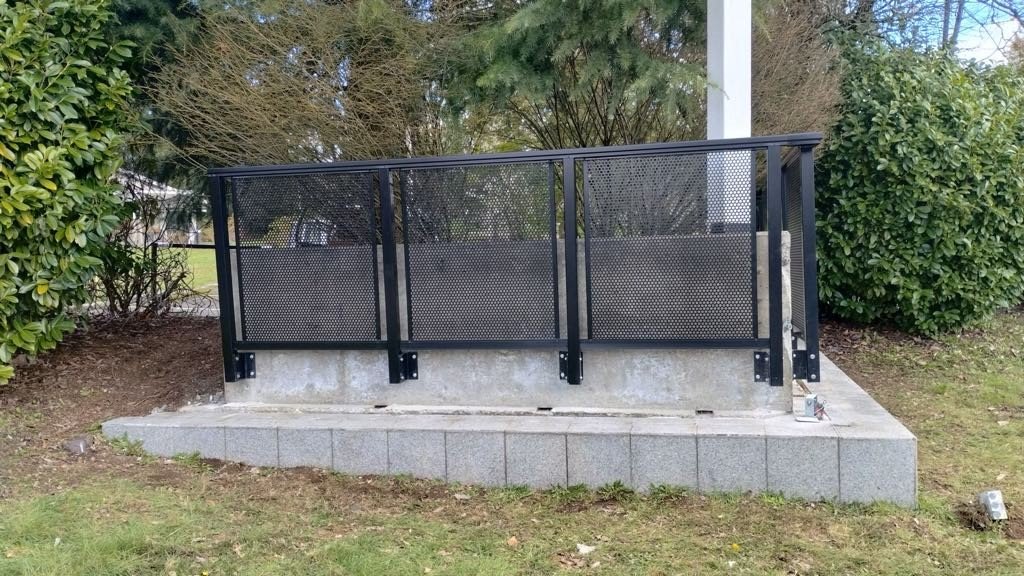 Outdoor black metal ramp with sides, installed on concrete foundation in a landscaped area with bushes and trees in the background.