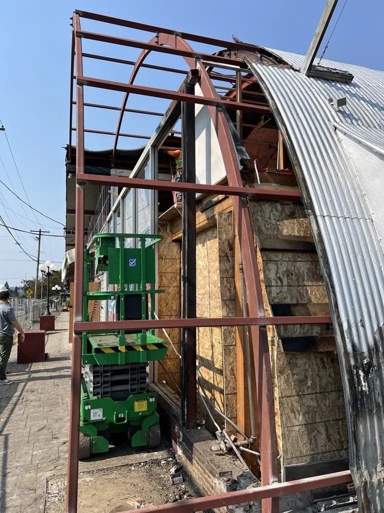 Construction site showing a partially built structure with a steel frame, plywood walls, and a corrugated metal roof. A green lift is positioned near the structure, and a person wearing a mask and a hat is standing nearby.