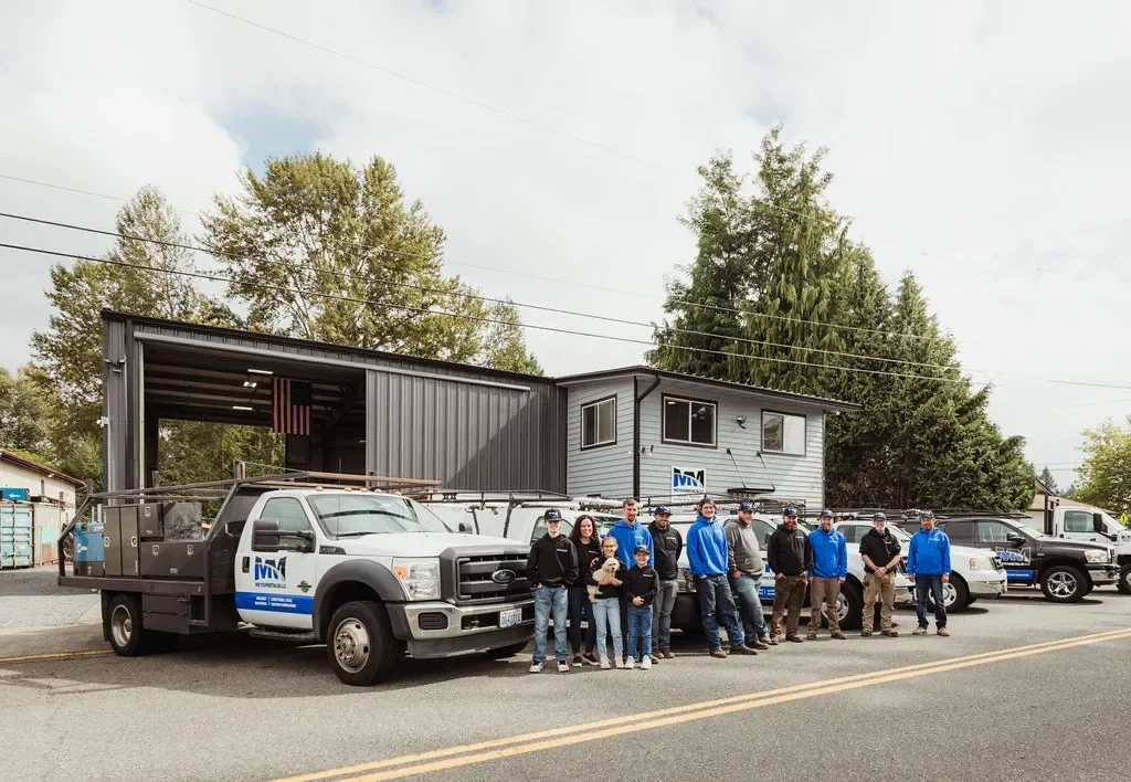 A group of people standing in front of utility trucks and a building, with trees and a cloudy sky in the background