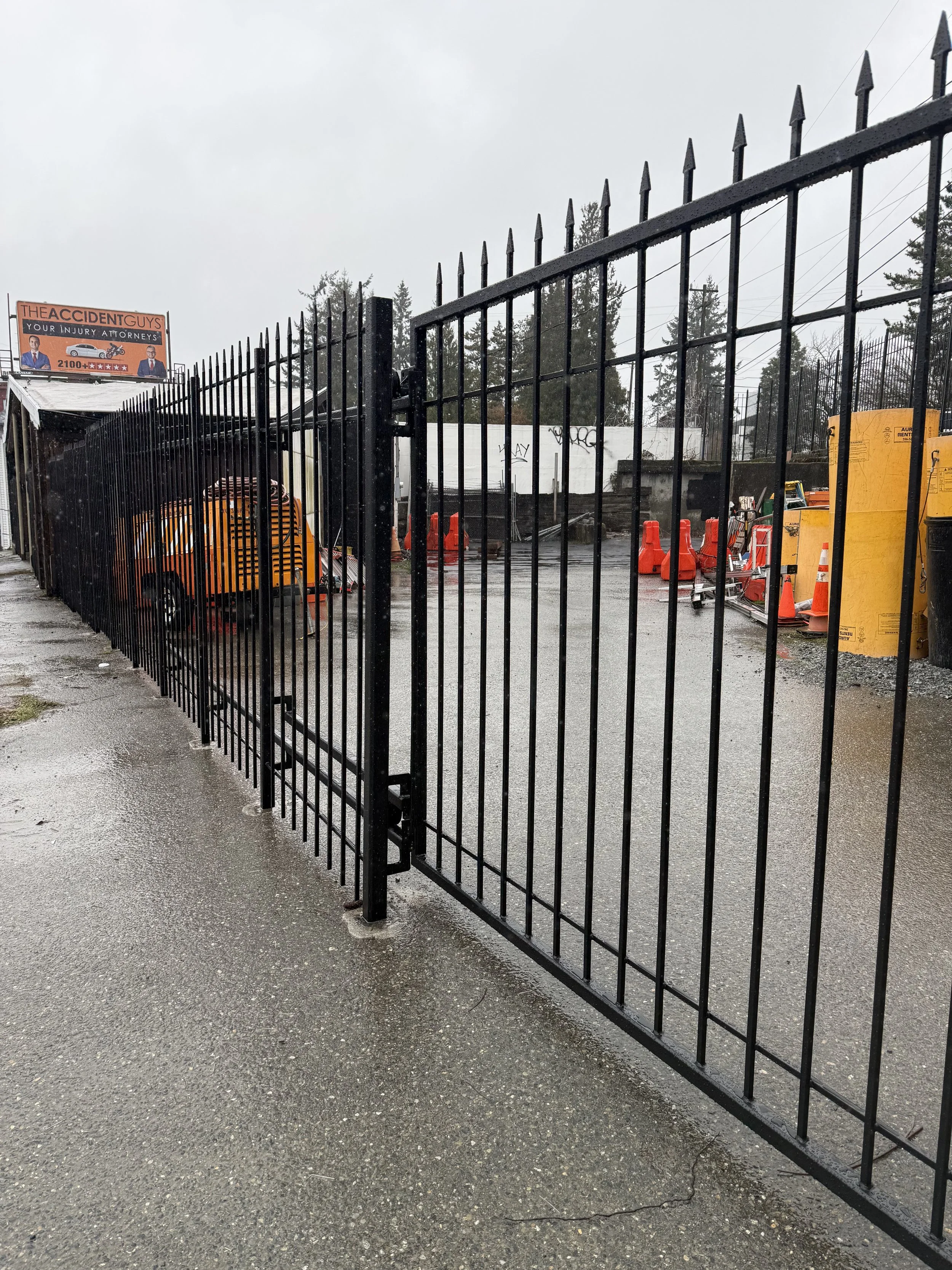 A black metal gate in front of a construction site or yard on a rainy day, with orange cones and construction equipment inside, and a sign for accident attorneys in the background.
