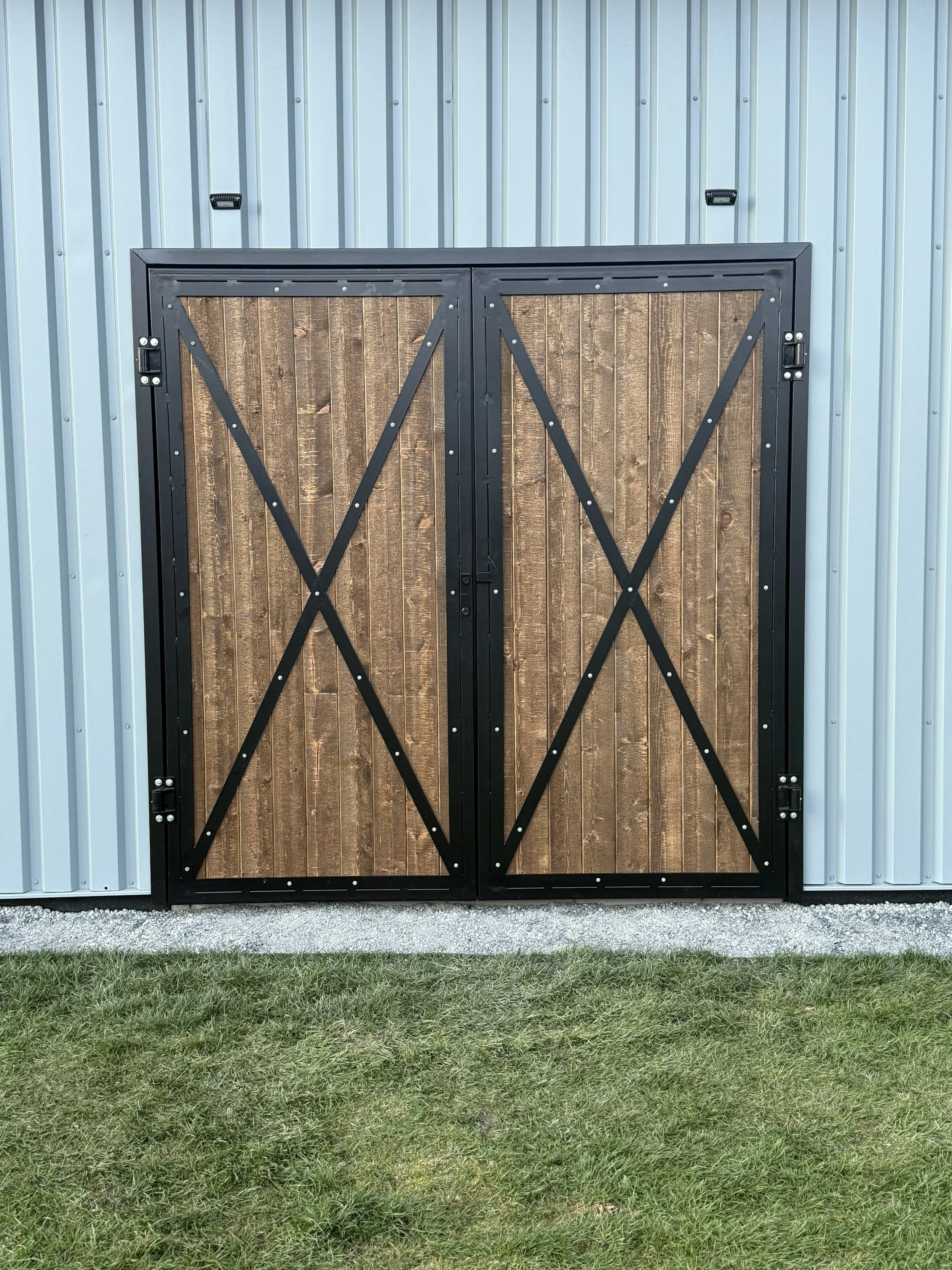 Double barn doors with wood panels and black metal frames, attached to a corrugated metal building, with grass and gravel in front.