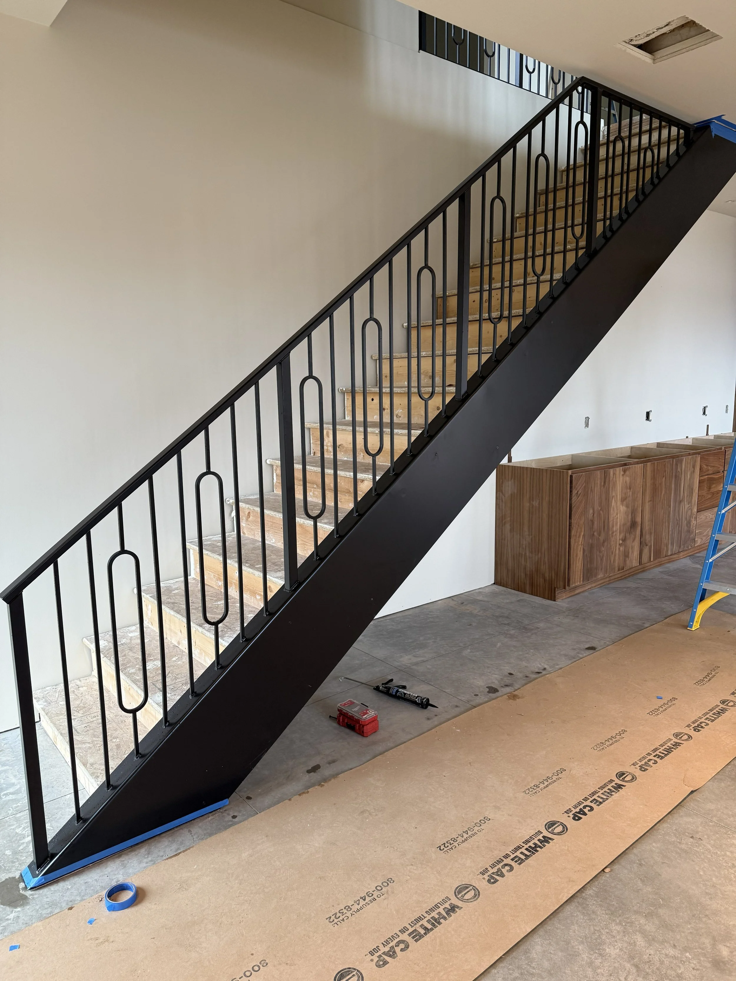 Interior of a building under construction with a black metal staircase, some tools and construction materials on the floor, and an unfinished ceiling with an opening.