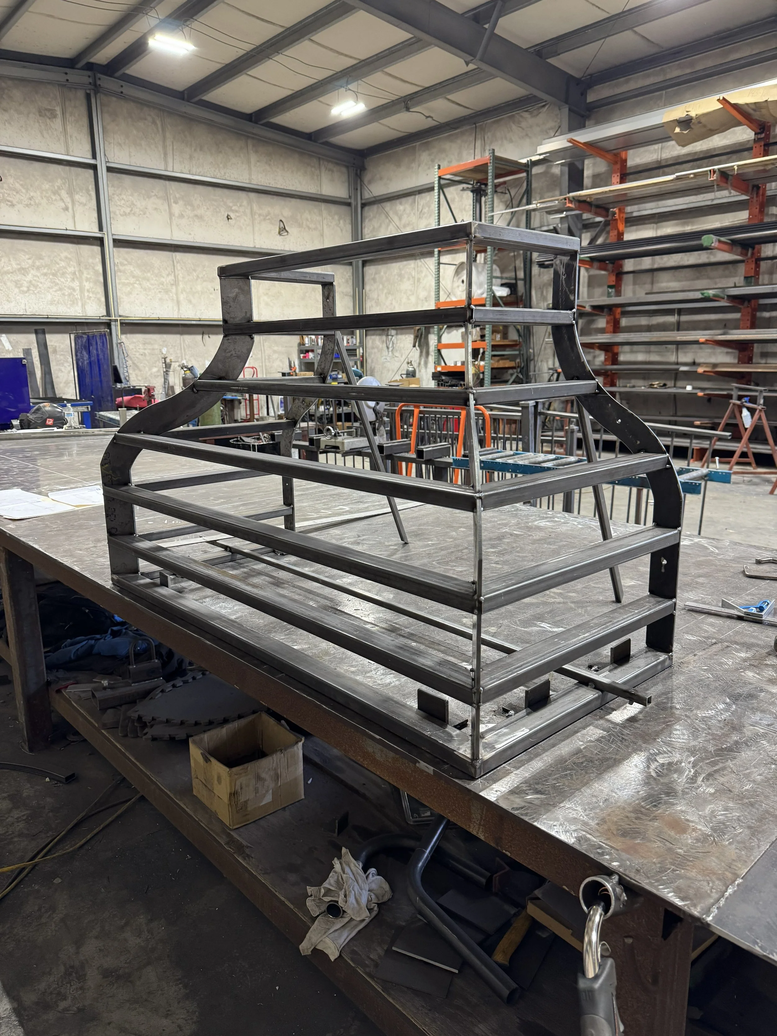 A metal frame structure being assembled on a worktable in a workshop, with shelves and tools visible in the background.