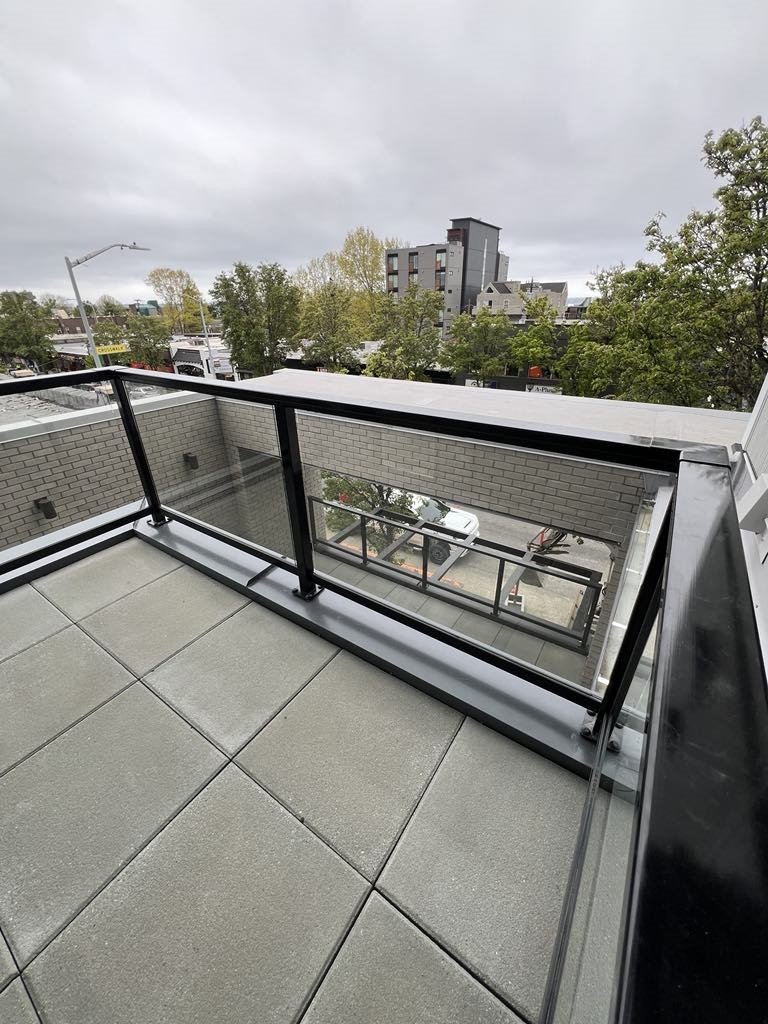 View from a balcony with tiled flooring and glass railing, overlooking trees, parked cars, and neighboring buildings under a cloudy sky.