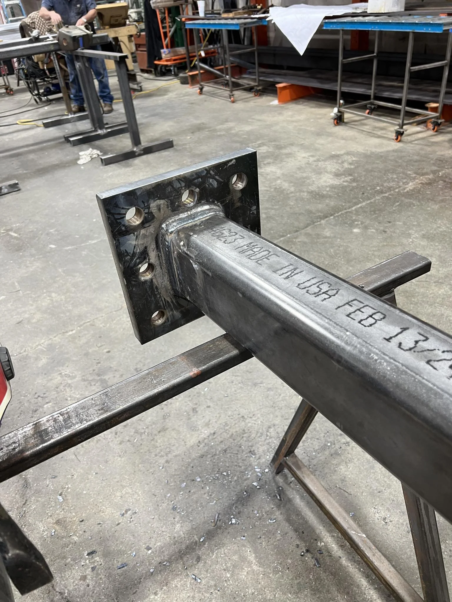 Close-up of a steel structural component on a worktable in a metalworking shop, with a workshop background including workbenches, carts, and tools.