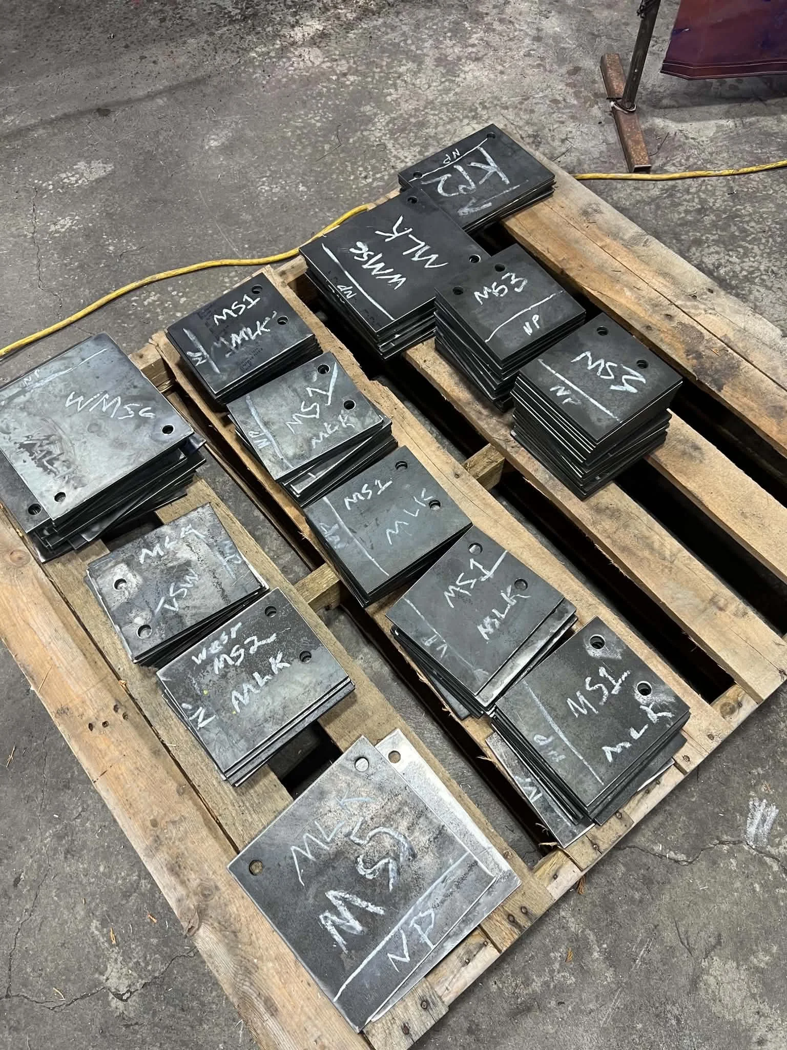 Metal plates labeled with chalk, stacked and placed on wooden pallets in a workshop setting.