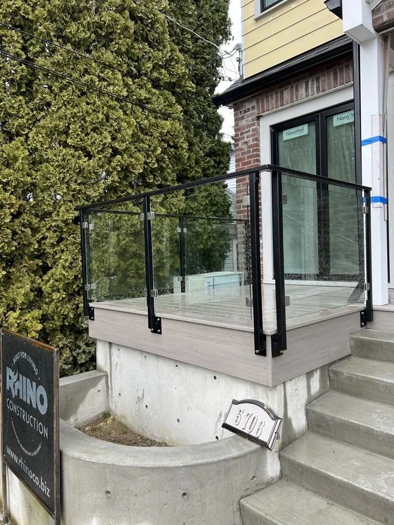 A small elevated balcony with glass railing, adjacent to a house with a brick wall and large window, steps leading up to it, and trees in the background.
