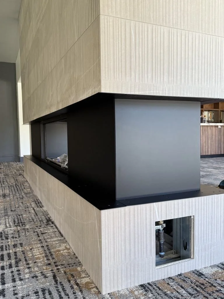 Partial view of a modern fireplace with a beige textured exterior, a black interior, and visible plumbing at the bottom, situated on a patterned carpet.