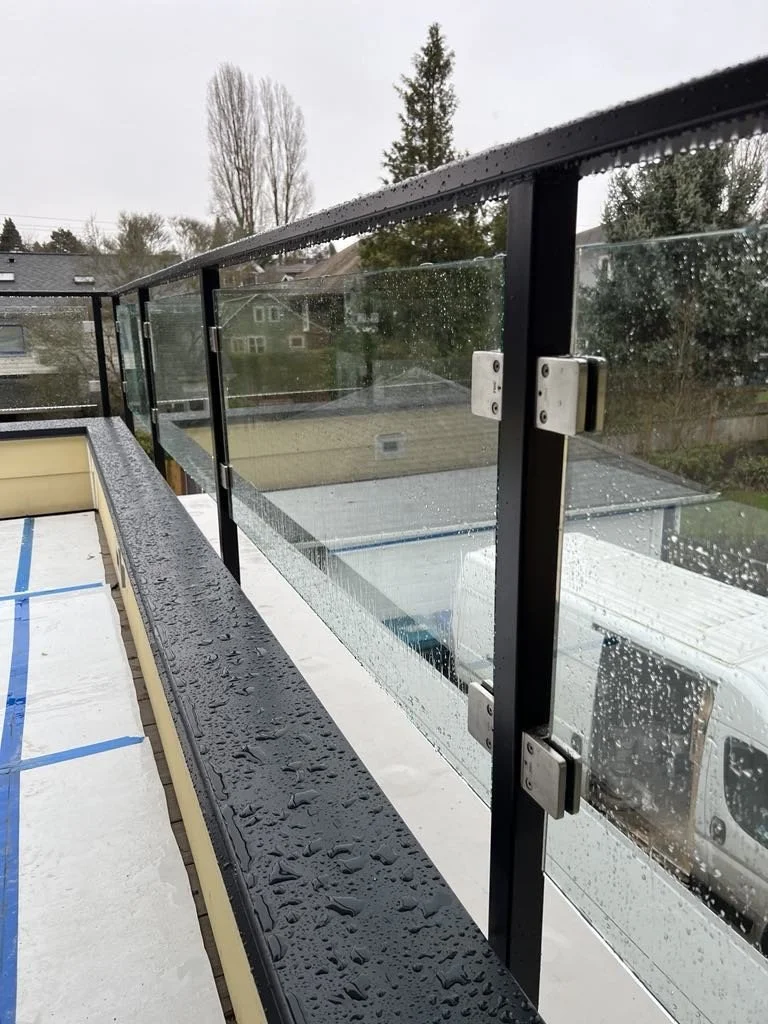 Rain drops on a black metal and glass balcony railing on a rainy day with residential houses and trees in the background.
