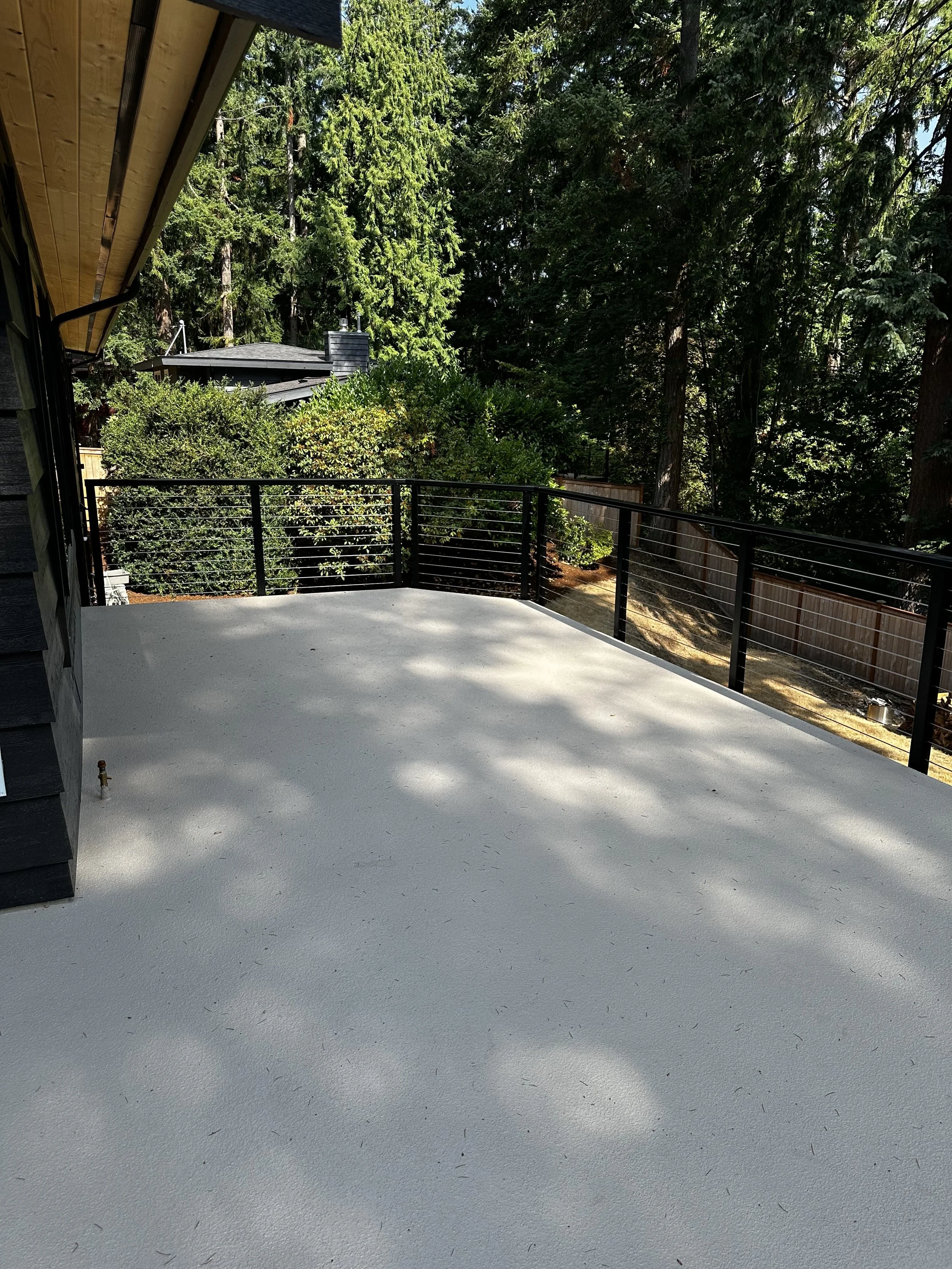 A view from a balcony or deck showing a white deck surface, black metal railing, and surrounding green trees and shrubbery.