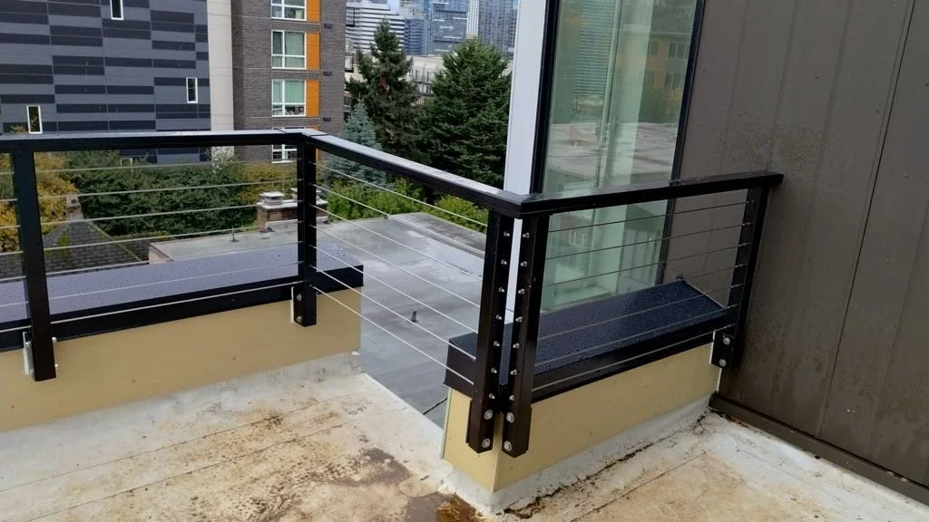 A balcony with a black metal railing and horizontal wires, overlooking an urban area with trees, rooftops, and modern buildings.