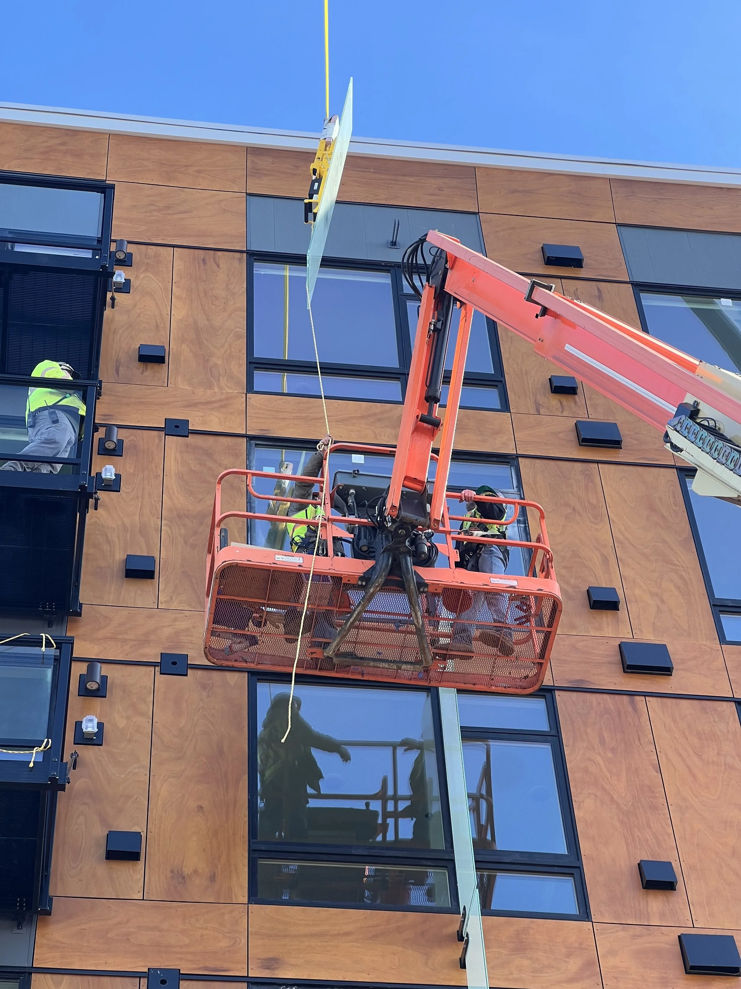 Workers in safety gear operate a lift to install a large glass panel on a building's exterior wall with wooden and glass panels.