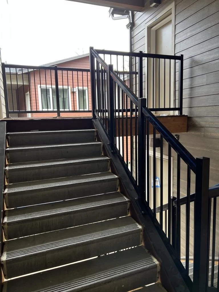 Outdoor metal staircase with black railings leading to an upper floor, next to a beige wooden wall with a white door.