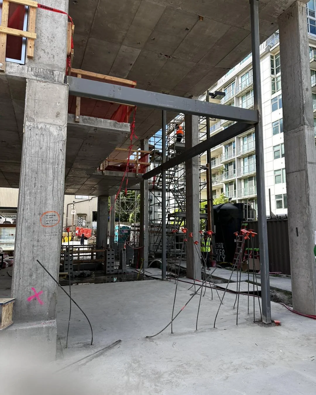 Construction site showing a partially built structure with concrete pillars, steel beams, scaffolding, and construction workers wearing safety vests and helmets in an urban area.