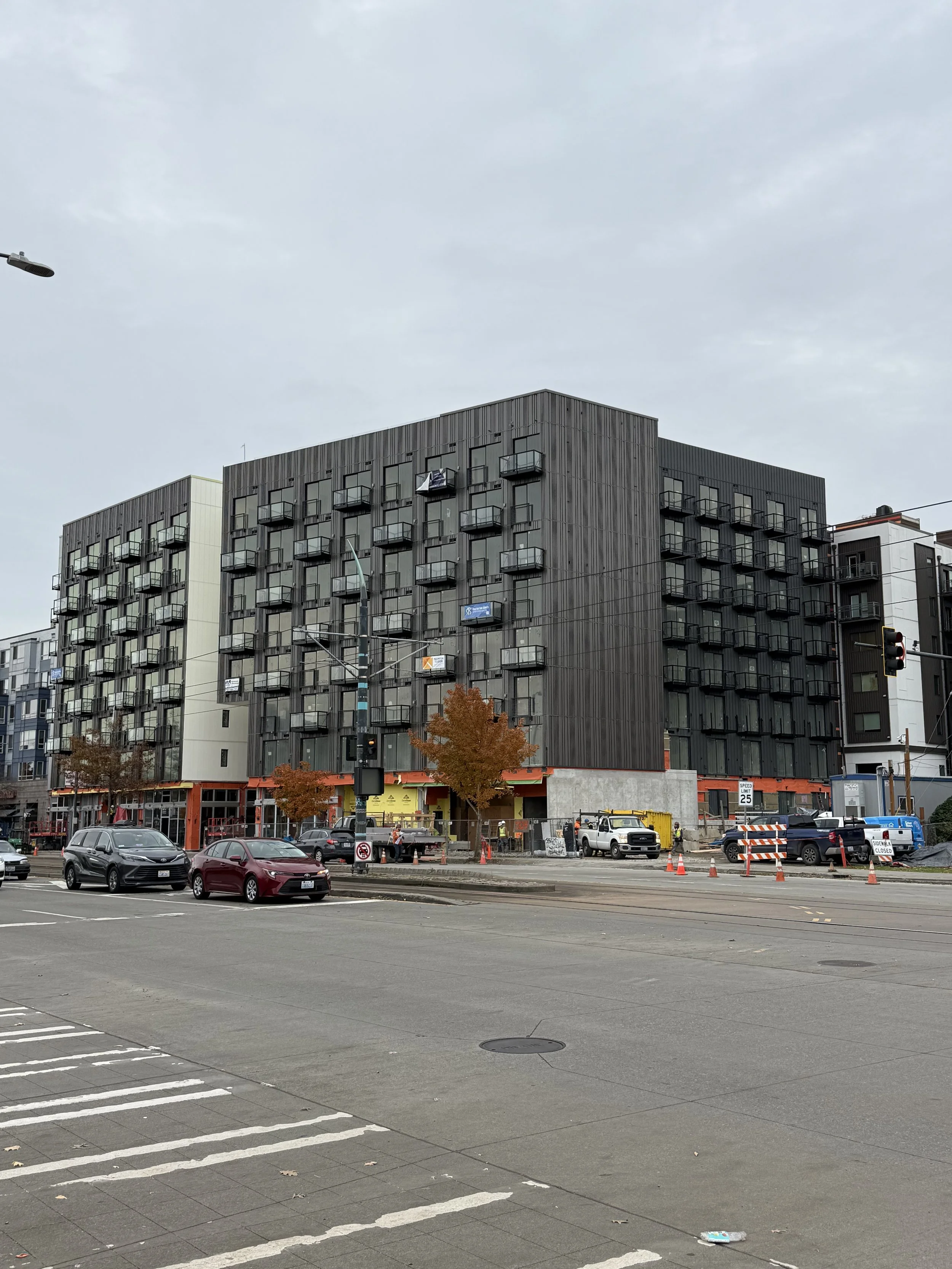 A city street scene with a multi-story residential building under construction, featuring black exterior walls and small balconies. There are several cars parked and driving along the street, and construction barriers and workers are visible near the building. The sky is overcast with gray clouds.