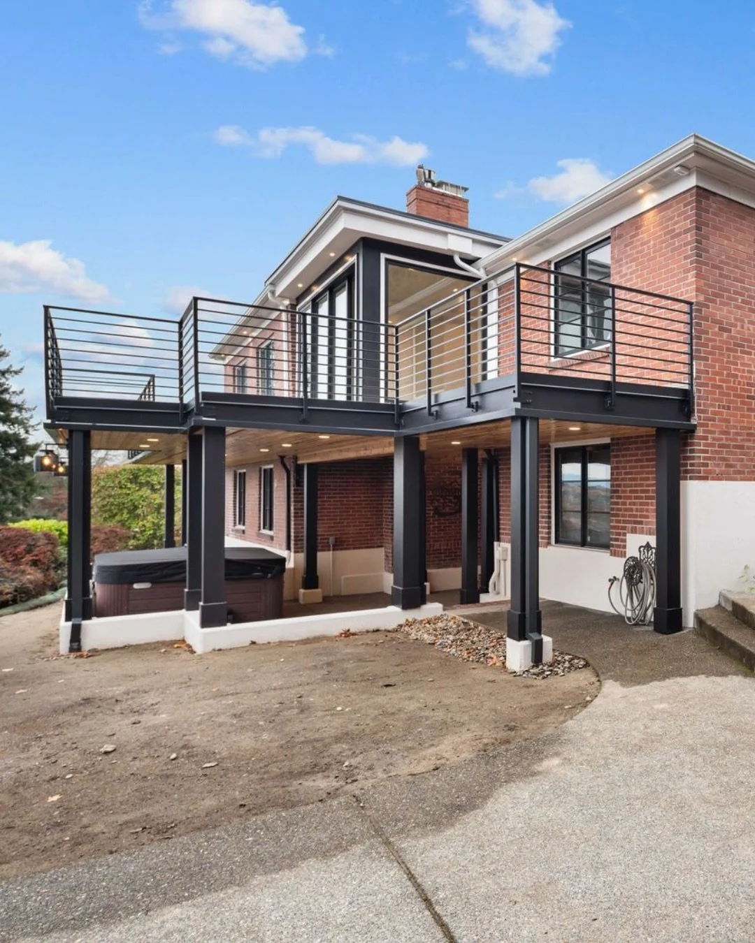 Modern two-story brick house with a black metal balcony and support columns in the backyard, with a hot tub on the ground level and a partly dirt yard surrounding it.