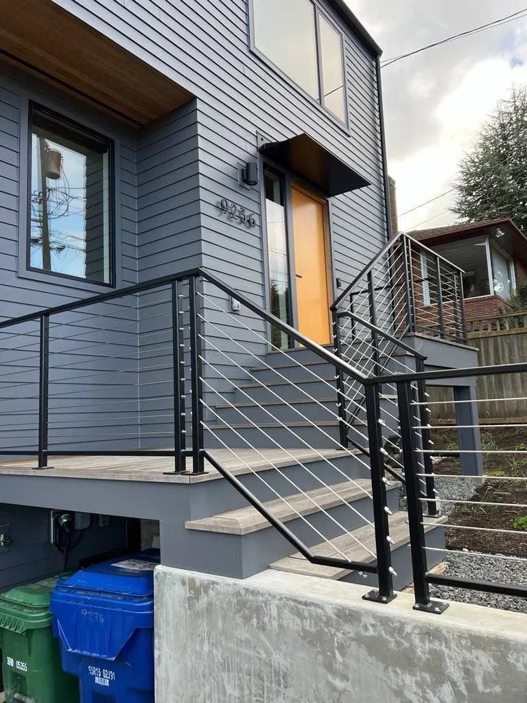Modern gray house with a black metal railing and a tan front door, front stairs, and a small porch. There are garbage bins at the bottom and surrounding fencing.