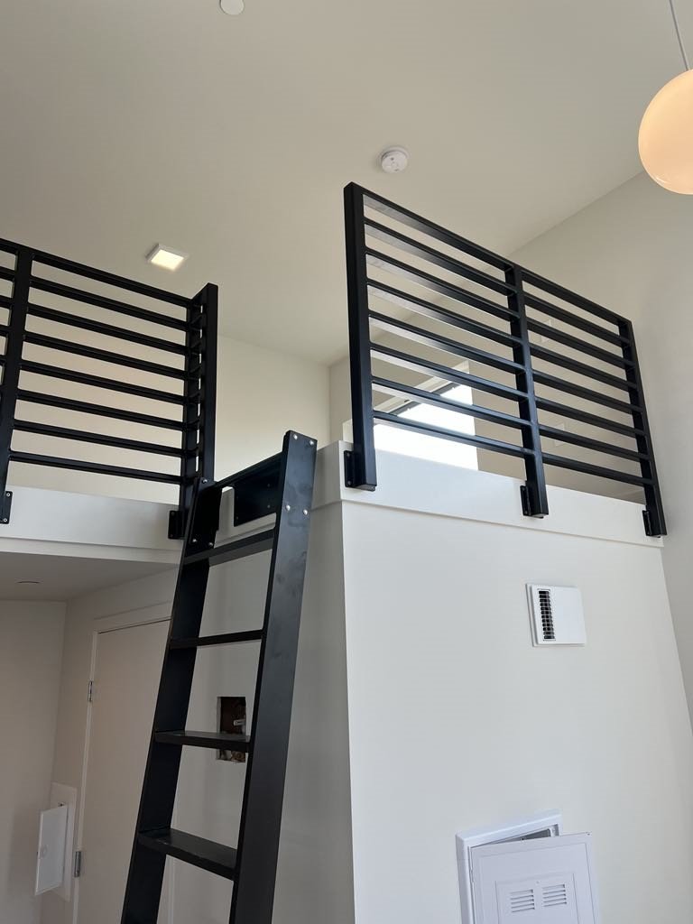 Interior view of a modern apartment with a black metal staircase leading to a loft area with a black metal railing. The ceiling is white with a square light, and there is a round pendant light. A vent and a thermostat are visible on the wall.
