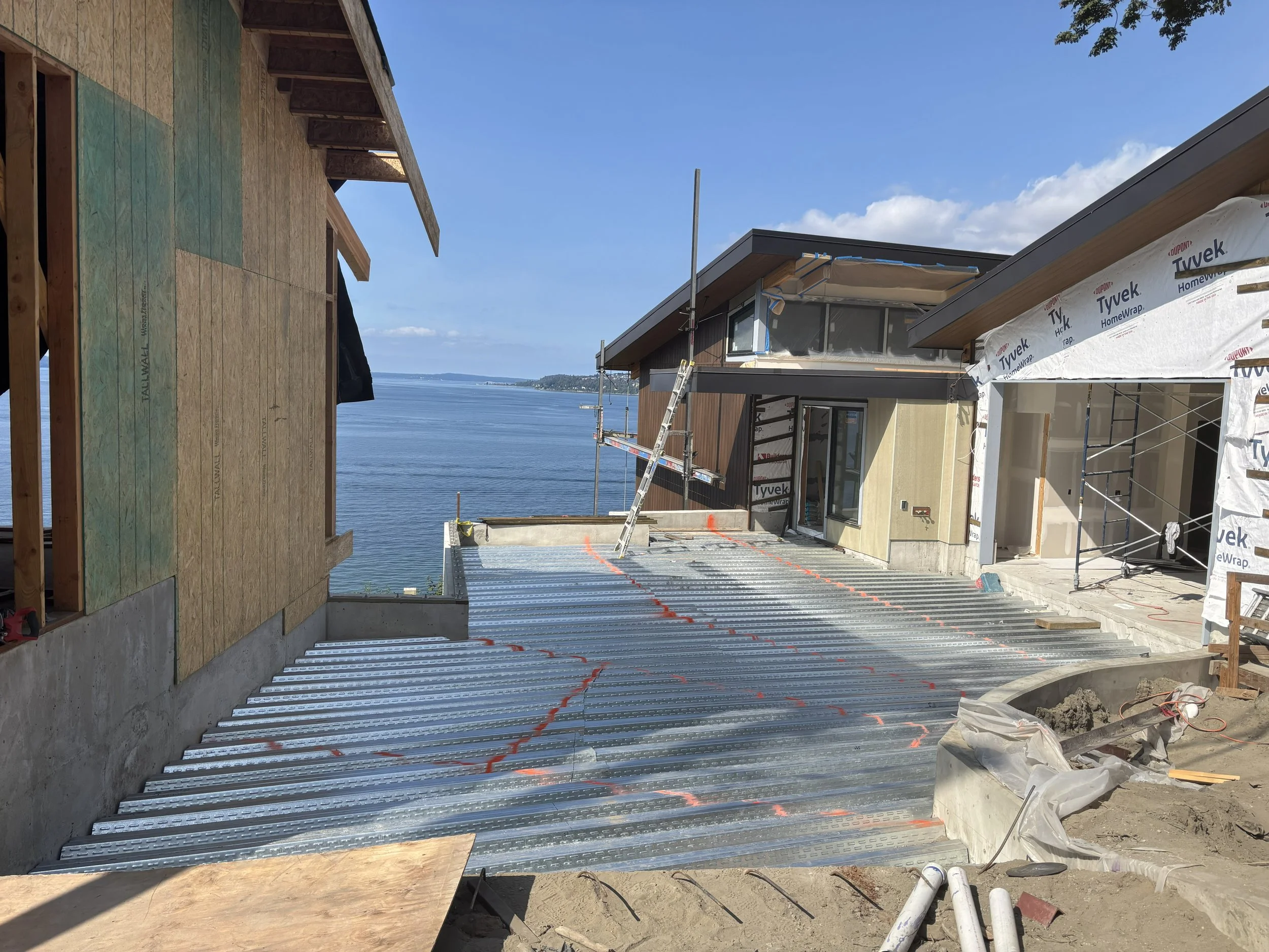 Construction site overlooking a body of water with buildings in the background, multiple houses being built with exposed wooden frames, scaffolding, and roofing in progress, with a clear blue sky.