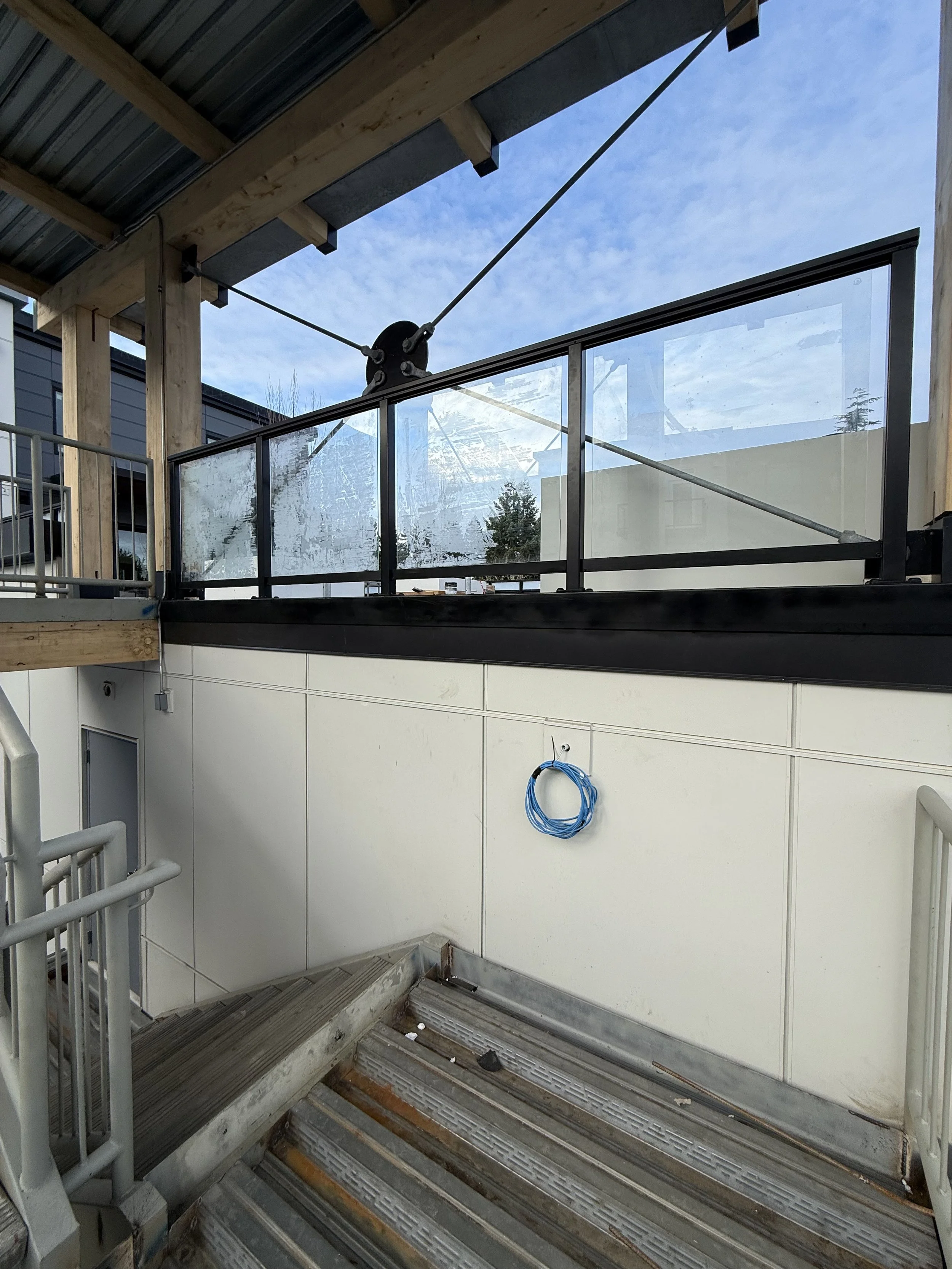 Construction site with metal stairs, a glass railing, wooden support beams, and a partly built structure. A coil of blue cable hangs on the white wall.