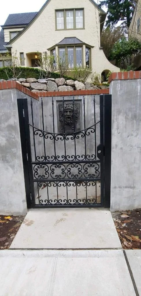 Black wrought iron gate with decorative swirls and a lion face emblem, set into a concrete and brick wall opening, leading to a garden and a white house with a bay window in the background.