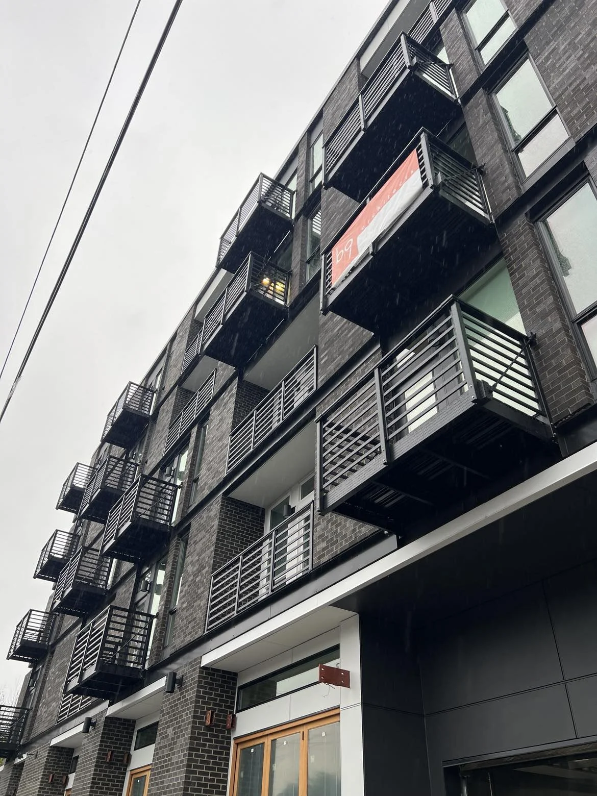 Modern multi-story apartment building with multiple balconies, some with metal railings, and visible windows. Overcast sky in the background.