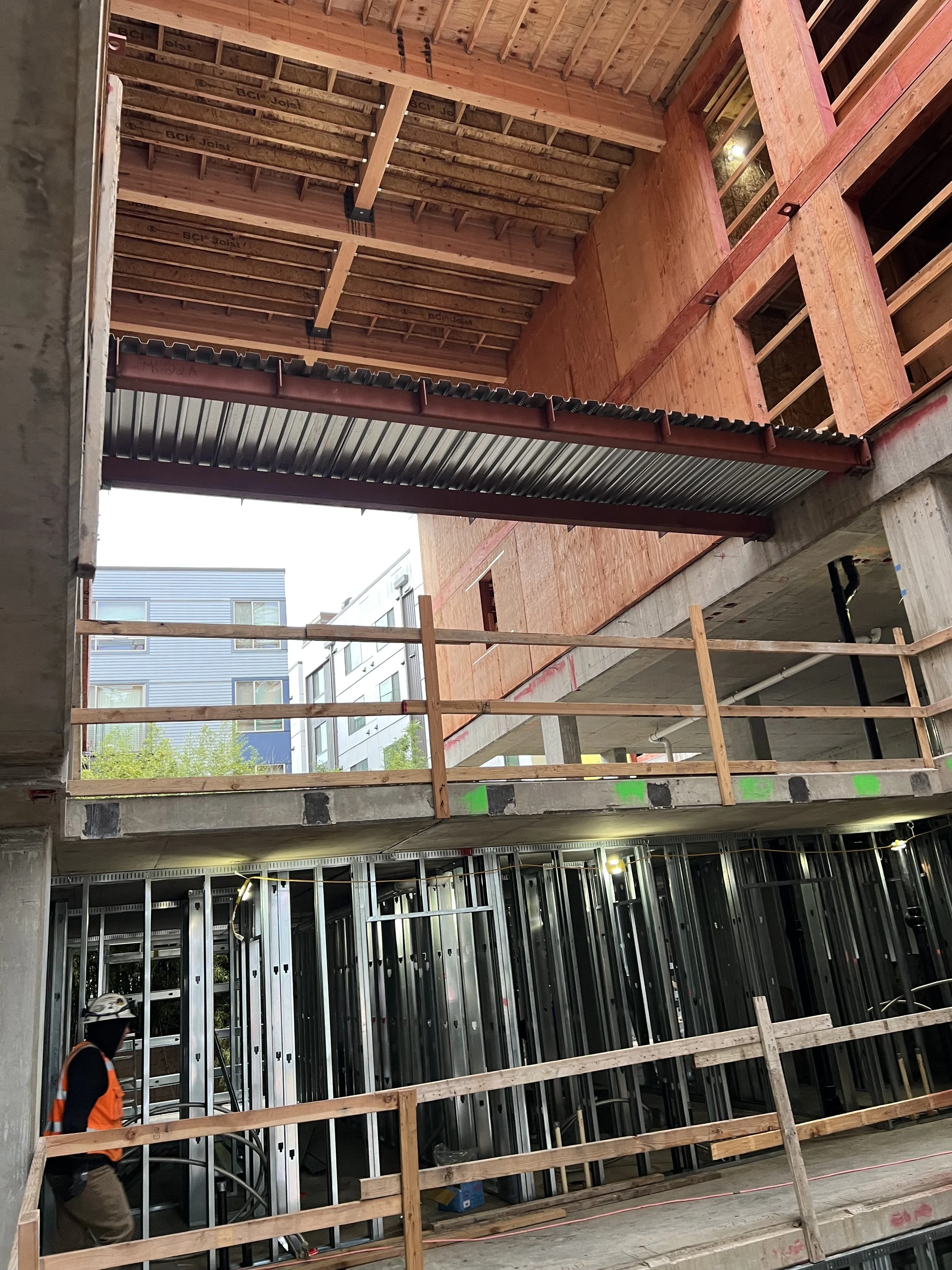 Construction site of a multi-story building with exposed wooden framing, steel beams, and workers in safety vests.