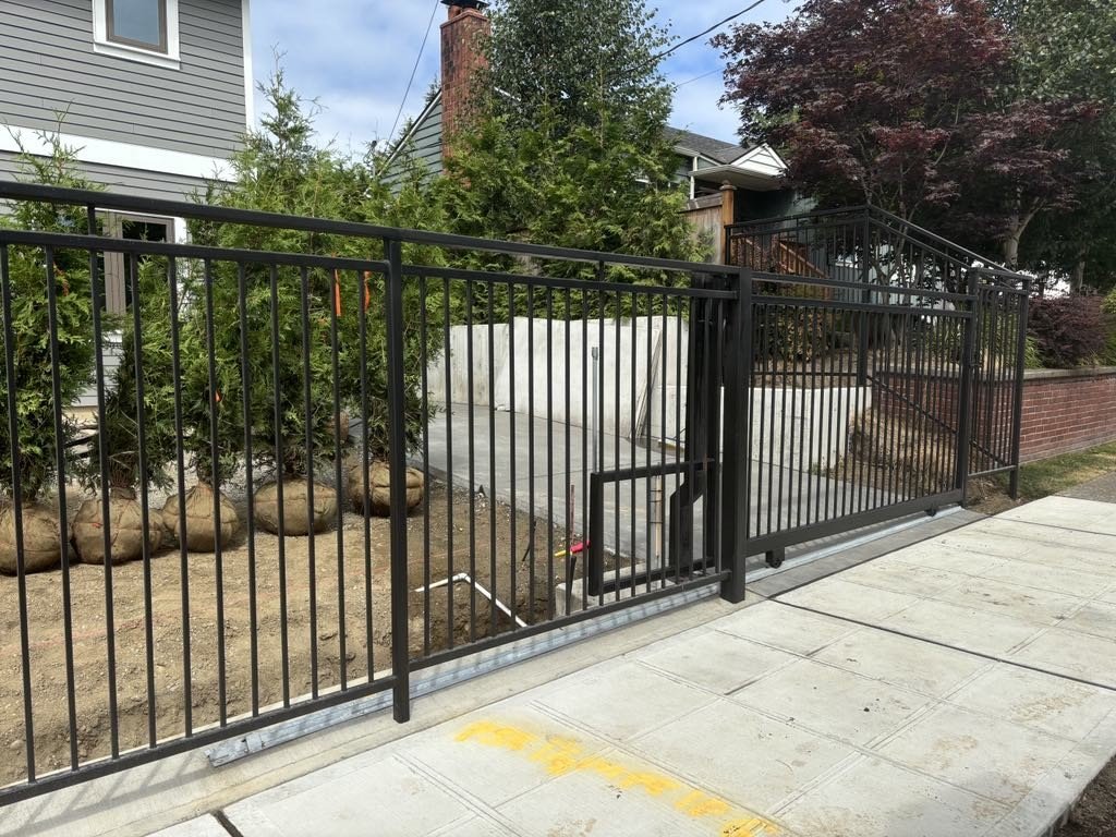 Black metal fence with gate enclosing a landscaped yard with a tree and shrubs, neighboring homes visible in a suburban setting.
