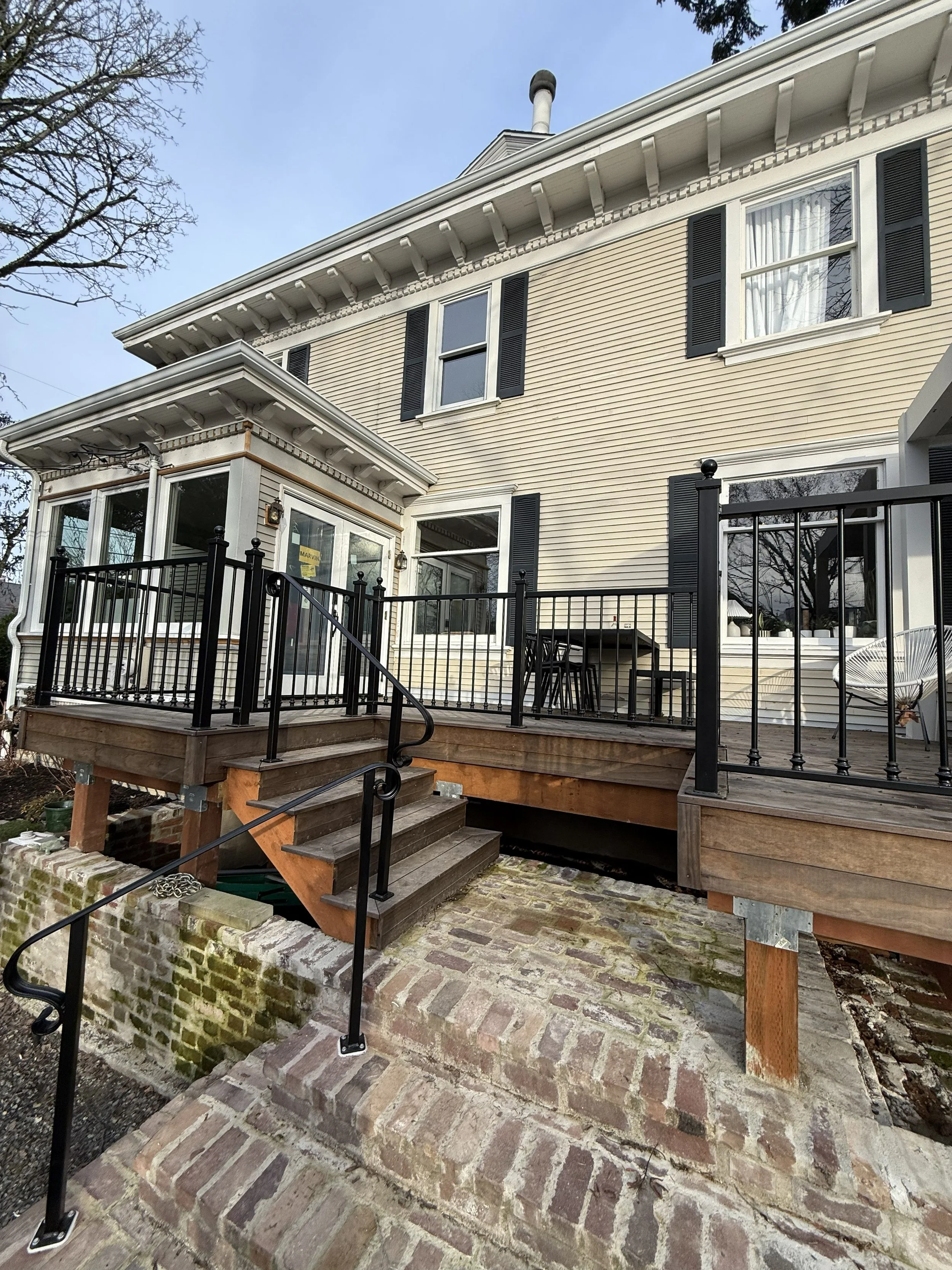 Back porch with wood steps and brick patio, black metal railing, and a screened-in porch extension, with a house with beige siding and white trim in the background.